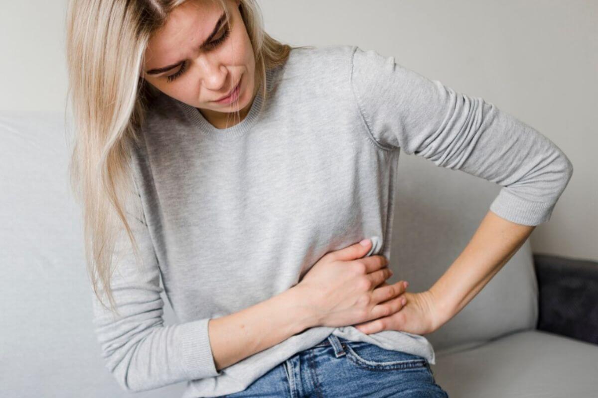 Fiatal szőke nő, szürke hosszú ujjú felsőben fájdalmasan fogja a derekát, oldalát.