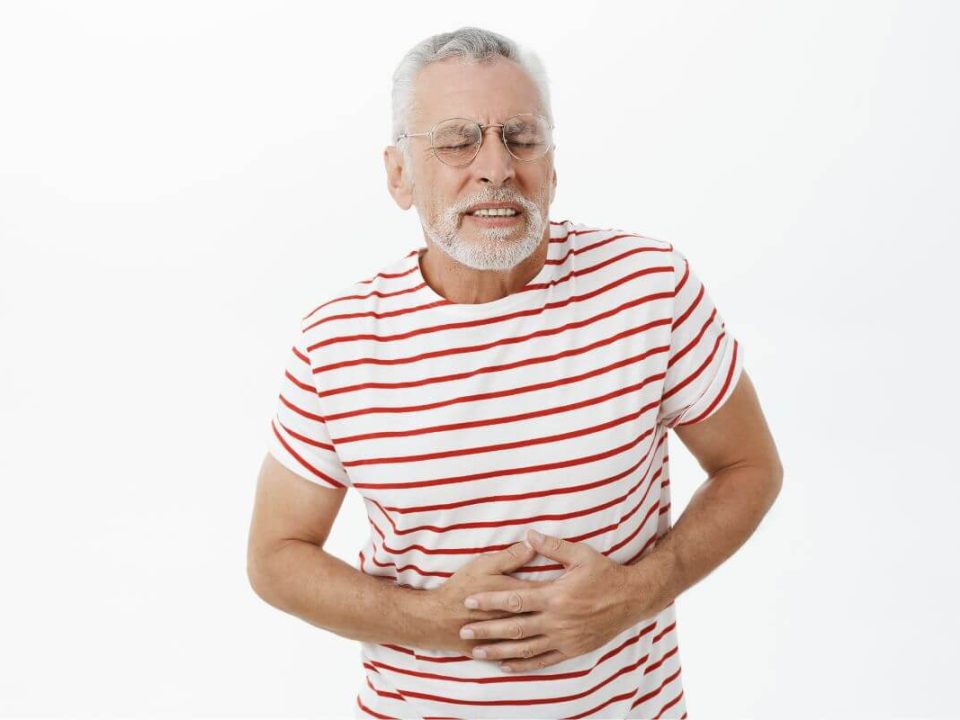 Fehér háttér előtt, szakállas, szemüveges, ősz hajú férfi, fehér-piros csíkos pólóban fájdalmasan fogja a hasát.