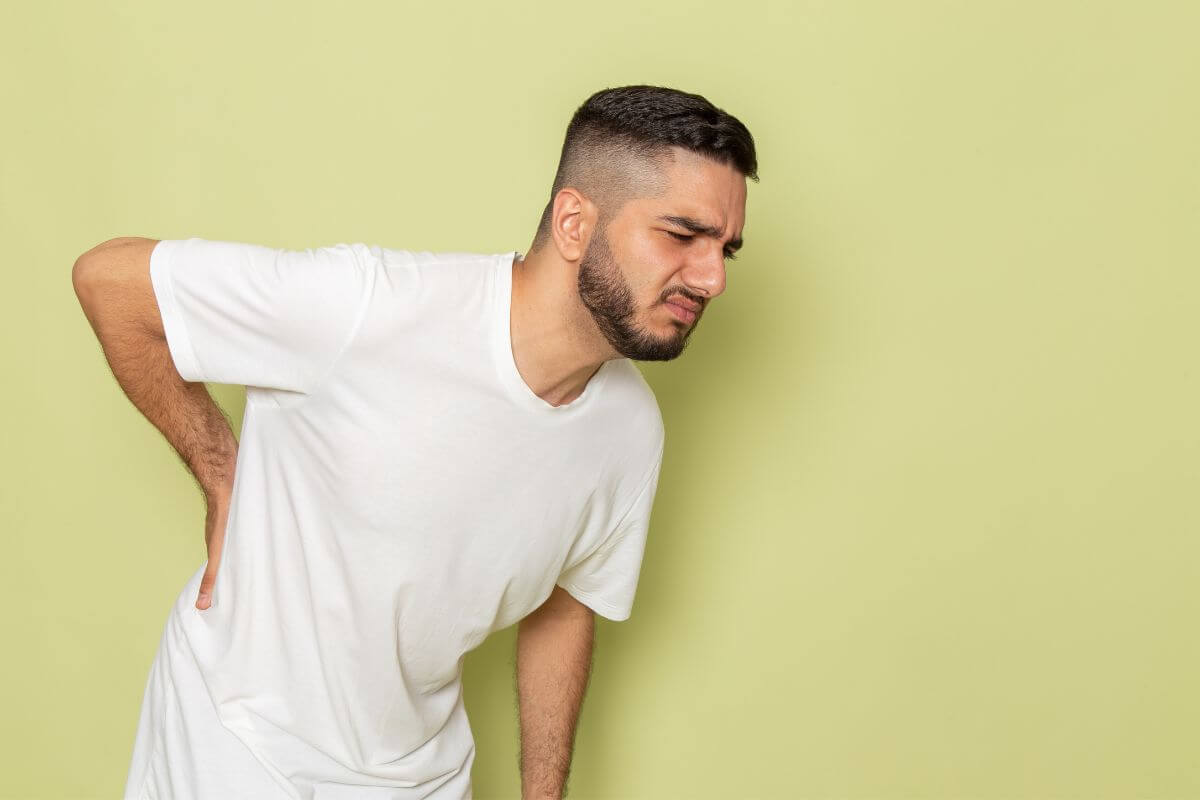 Fiatal, hosszú barna hajú lány, fehér ingben és farmerban a derekát fogja fájdalmasan. Egy irodában van, előtte az asztalon egy monitor és egy kávéspohár látszik.